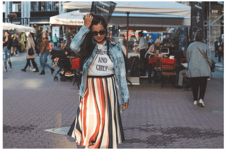 A woman wearing a short sleeve graphic tees with stripe skirt & denim jacket walking through a fair looking very trendy & feeling great on her body image.