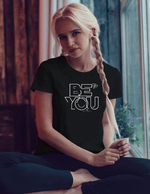 A blonde woman wearing a black inspirational t-shirt with white graphics Be You sitting down in front of a window