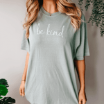 woman in a standing wearing a positive t shirt in green bay color - screen says Be Kind