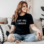 You Are Enough Motivational Black Short Sleeve Shirt (Unisex) in black color with white letter on model sitting down with legs crossed over one another.