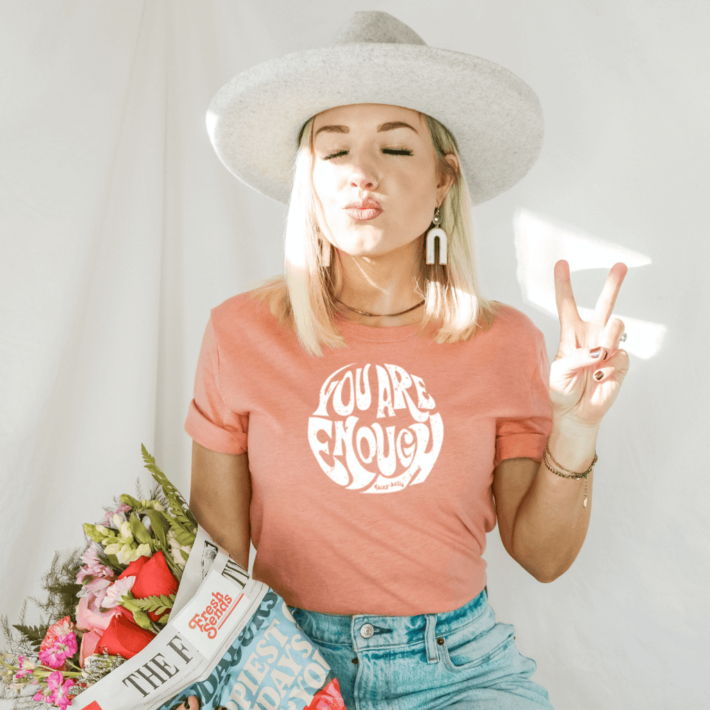 Women in a hat wearing an inspirational tee in sunset peach color screen says you are enough